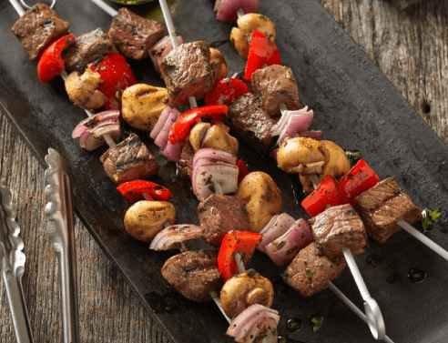 Beef kabobs on a skewer, mixed with chunks of red bell peppers, mushrooms, and red onions, arranged on a dark slate serving tray